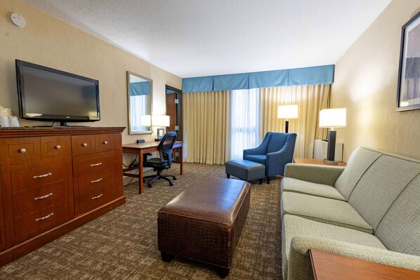A hotel room with a flat-screen TV, a wooden dresser, a desk with a chair, a blue armchair, a sofa, and a large window with curtains. - Pear Tree Inn St. Louis Near Union Station (St. Louis)