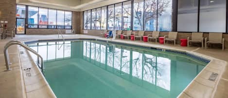 An indoor swimming pool with clear glass walls, a tiled deck, and a row of chairs along the poolside.