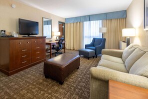 A hotel room with a wooden dresser, a television, a desk with a chair, a blue armchair, a beige sofa, and a patterned carpet.