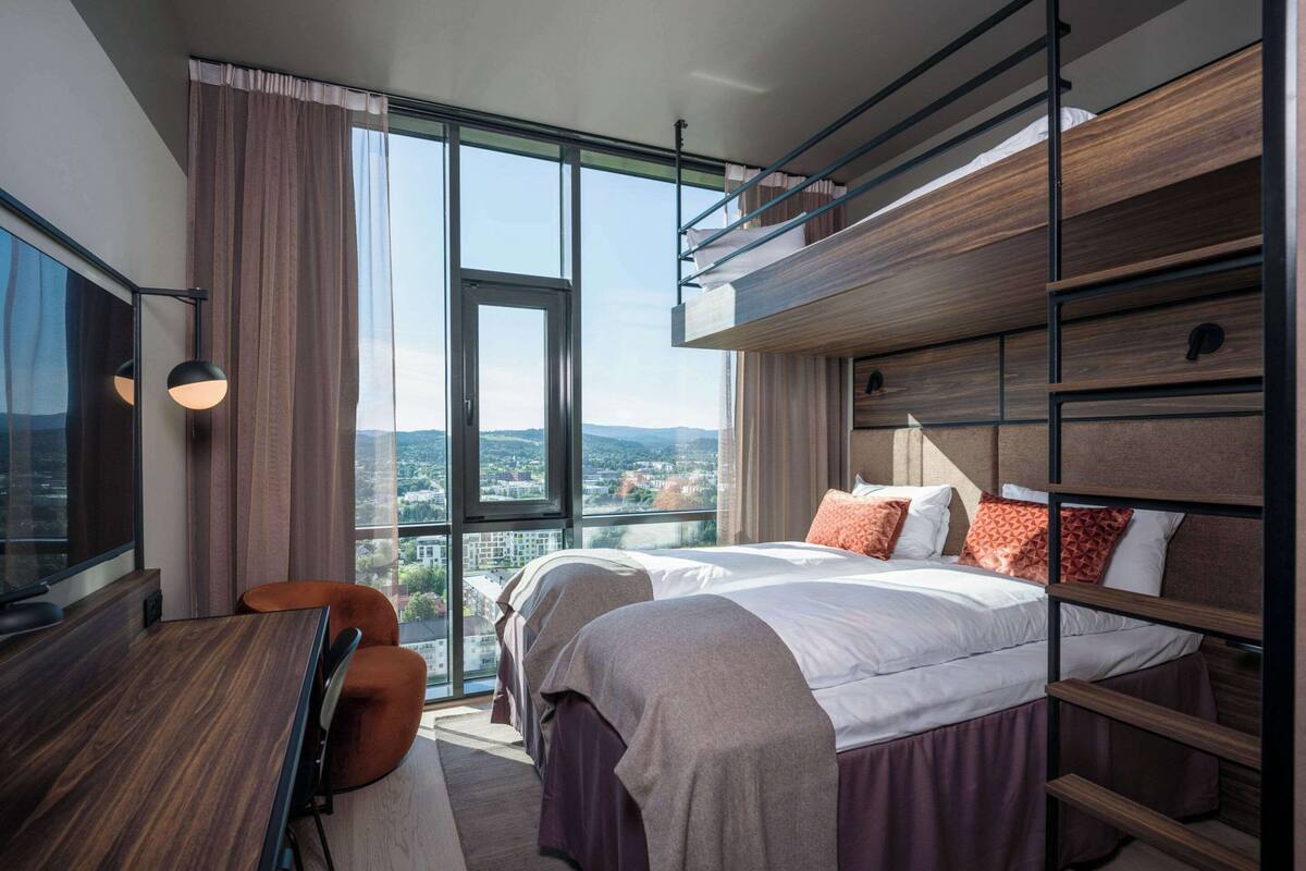 A modern hotel room with a large window, a desk, two beds, and a view of the city.