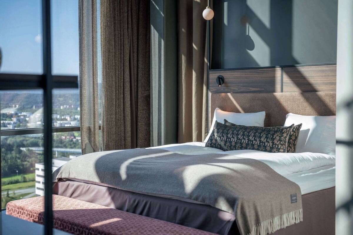 A modern bedroom with a large bed, a bench at the foot of the bed, and a city view through large windows.
