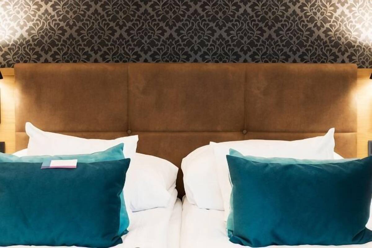 A bed with a brown headboard, white and teal pillows, and a patterned wallpaper background.