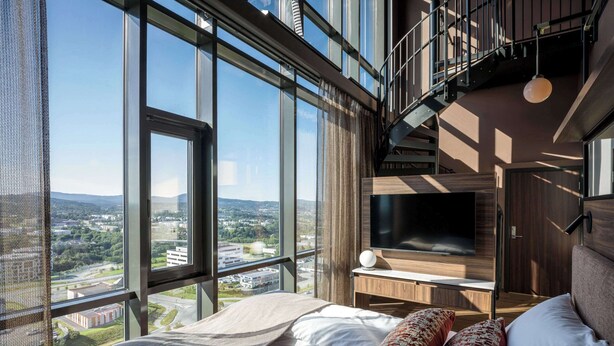 Un dormitorio moderno con una cama grande, un televisor y vistas de la ciudad a través de amplios ventanales.