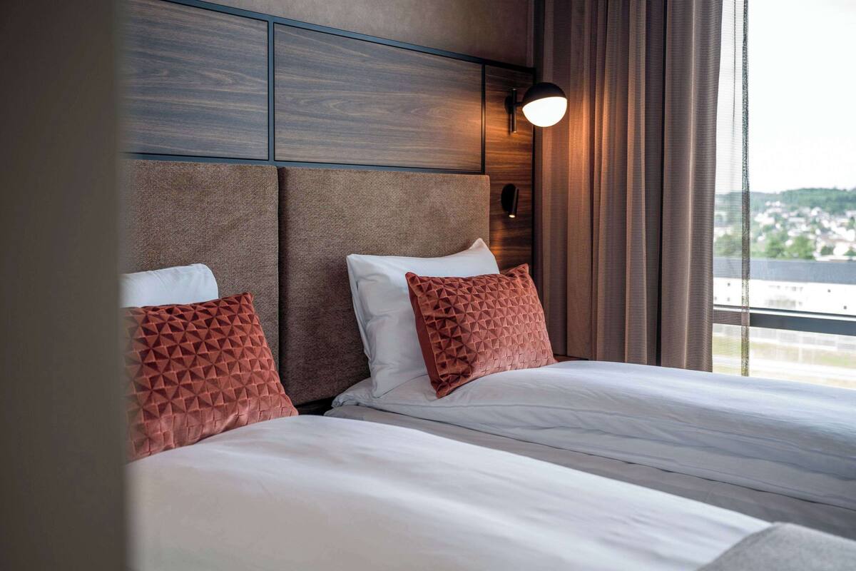 A hotel room with a large bed, two pillows, a headboard, a wall-mounted light, and a view through a window.
