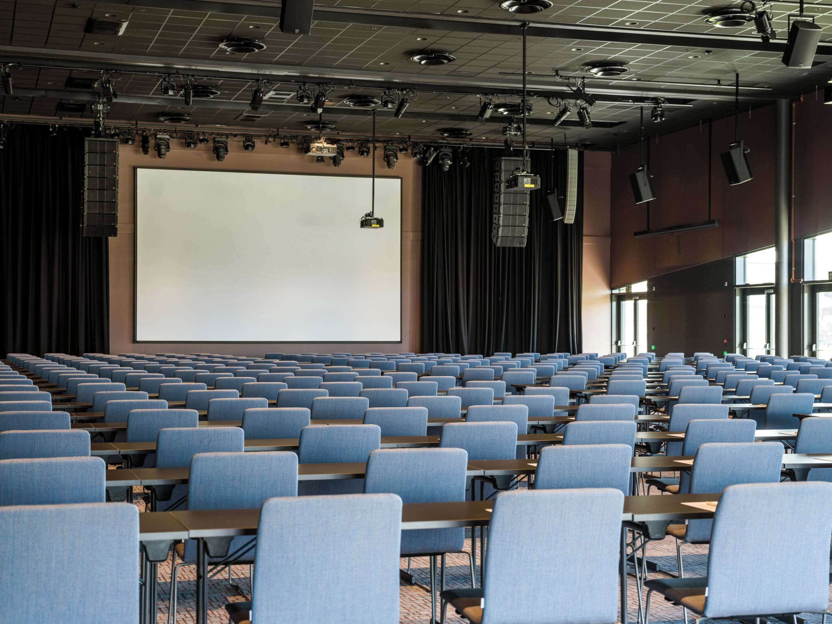 A large auditorium with rows of blue chairs and tables, a large screen, and overhead lighting.