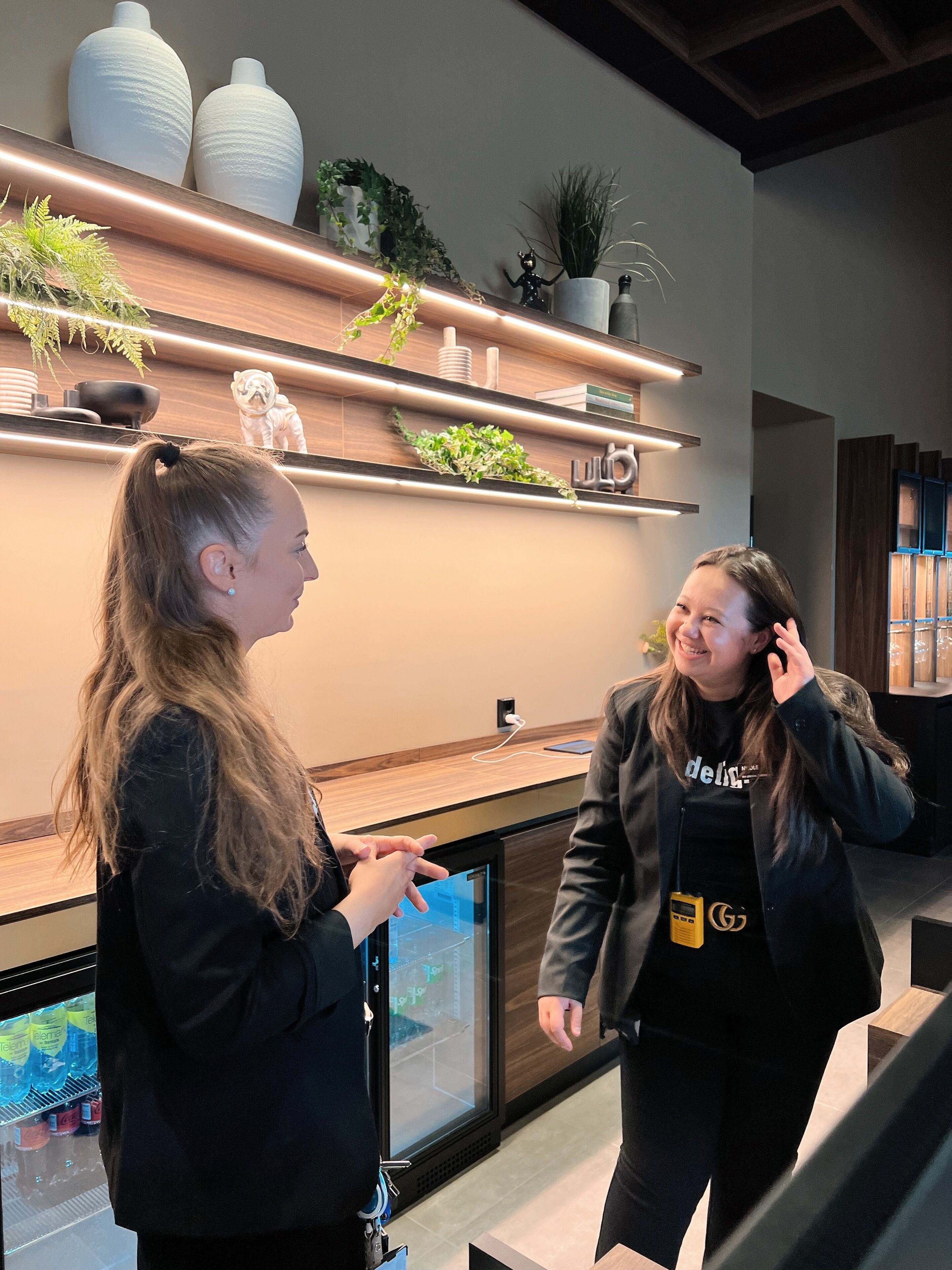 Dos personas conversando en un interior moderno con estanterías de madera, plantas y una pantalla digital.