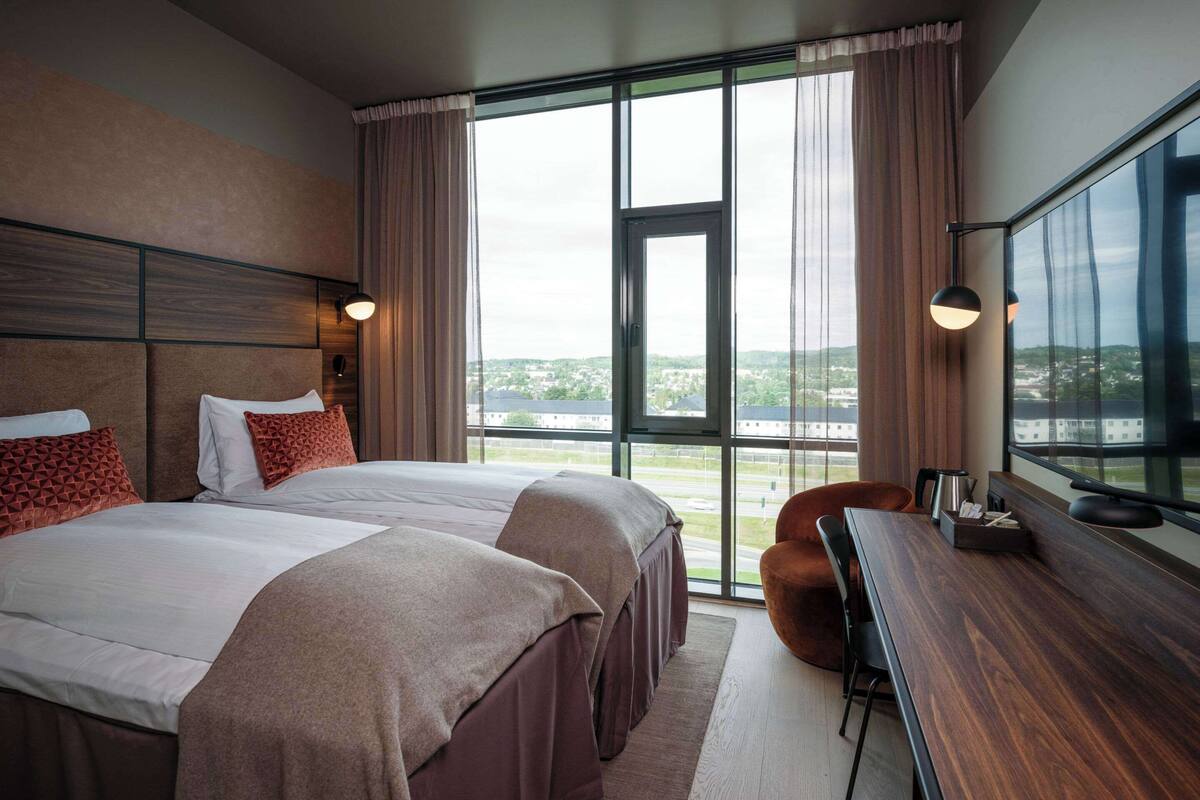 A hotel room with two beds, a desk, and a large window offering a city view.