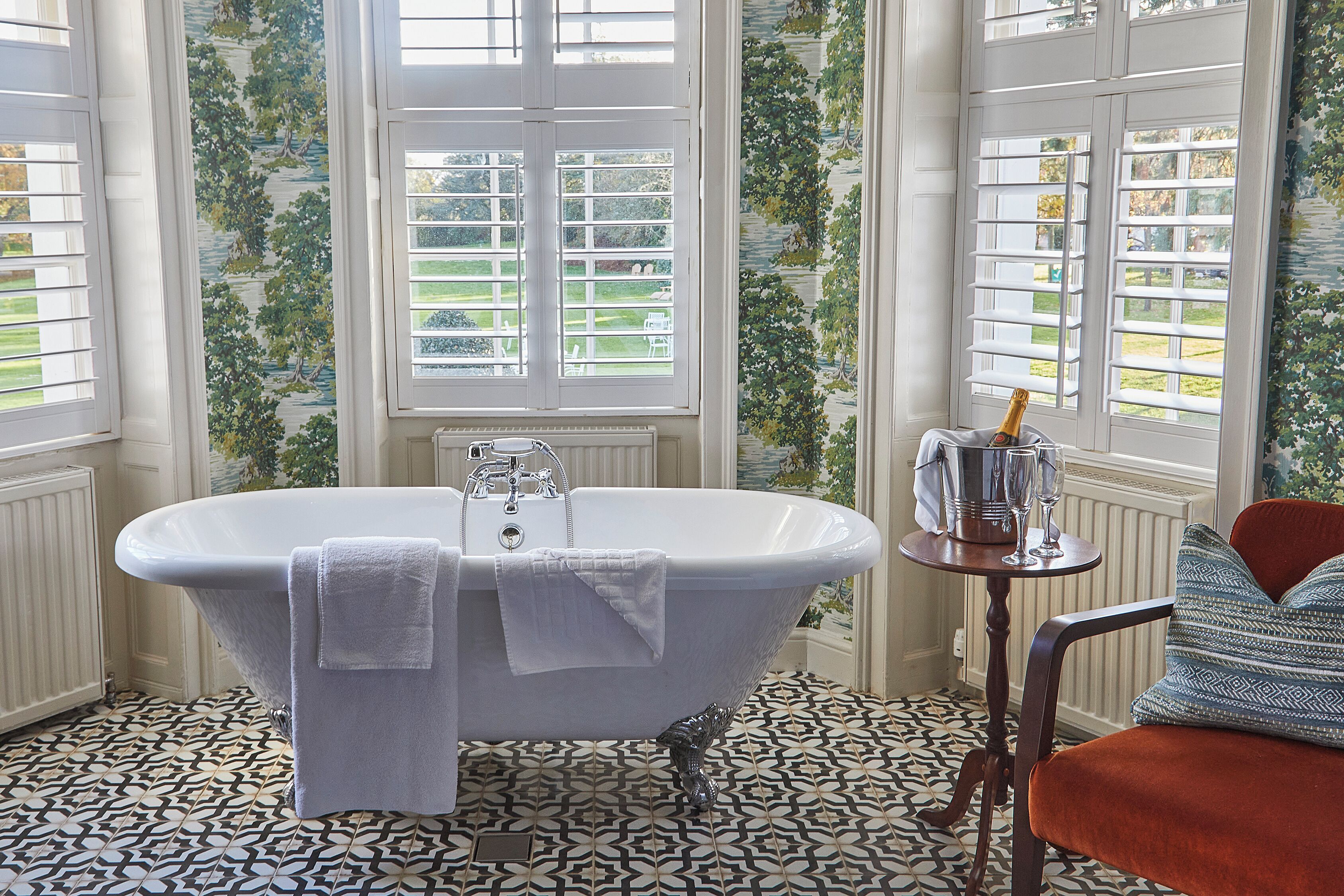 A bathroom with a freestanding bathtub, a radiator, a small table with a bottle and glasses, and a chair with a cushion.