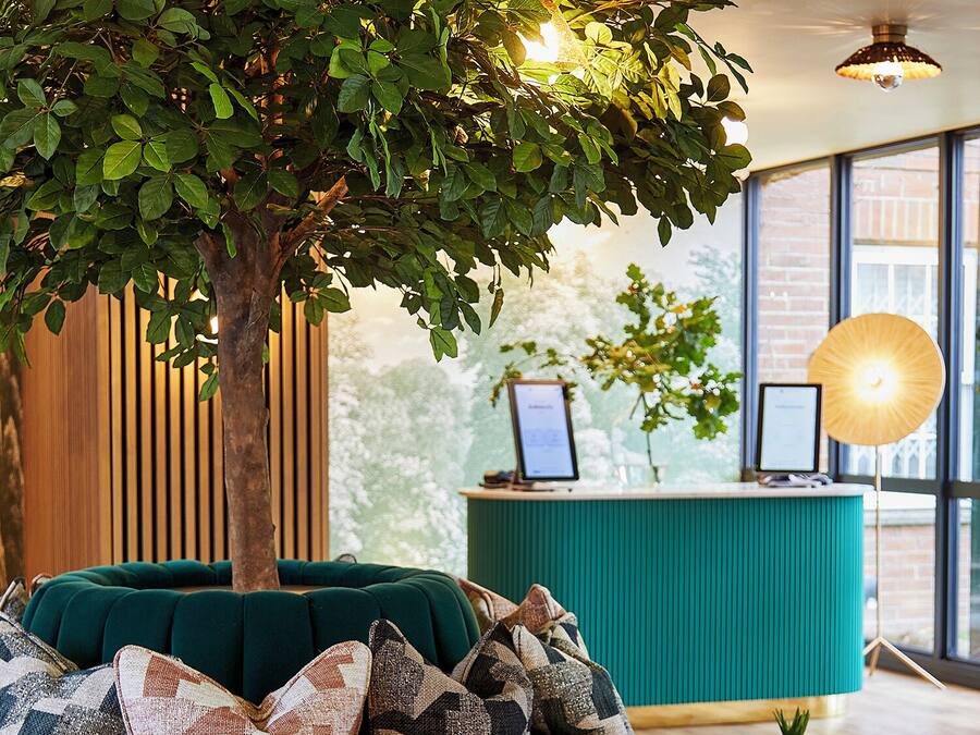 A teal sofa with patterned cushions, a small table, and a tree in a modern lobby with wooden slats and a reception desk.