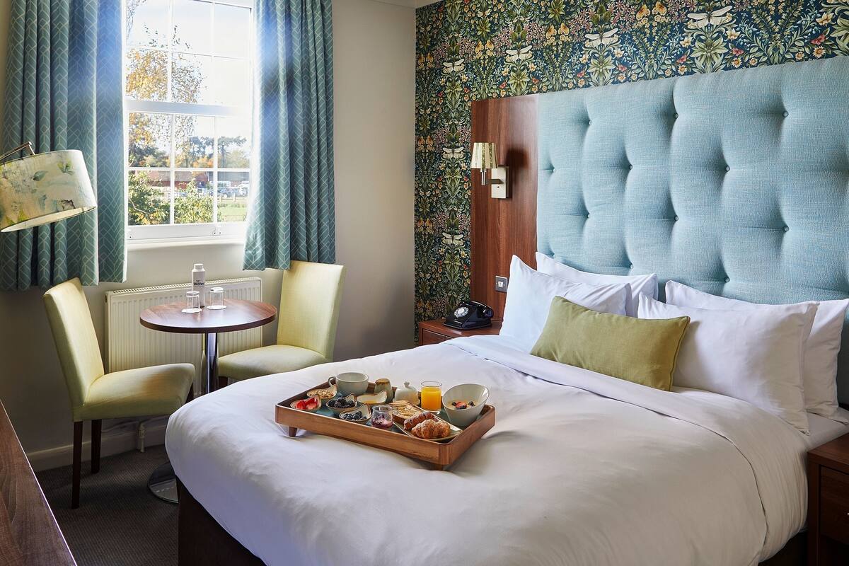 Quarto de hotel com uma cama grande, bandeja de café da manhã, duas cadeiras, uma mesa pequena e uma janela com cortinas.