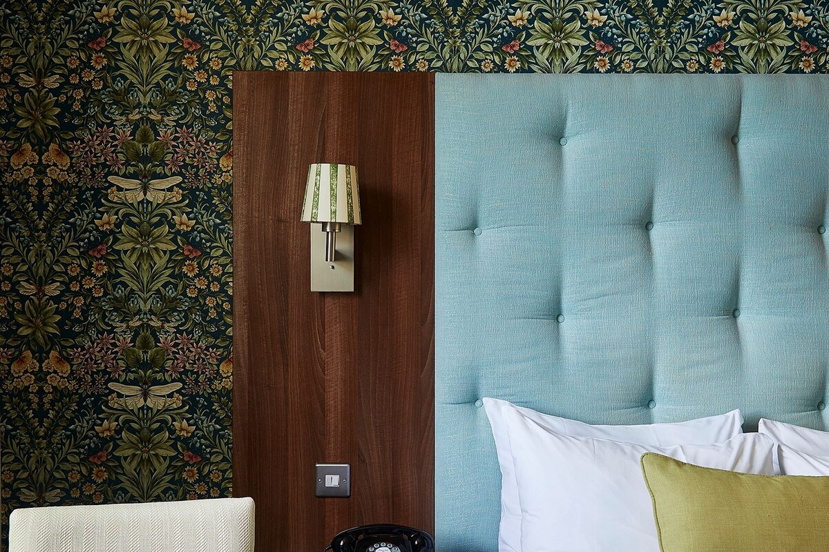 Una habitación de hotel con papel tapiz floral, una mesita de noche de madera, una mesilla con una botella de agua, vasos y un teléfono giratorio.