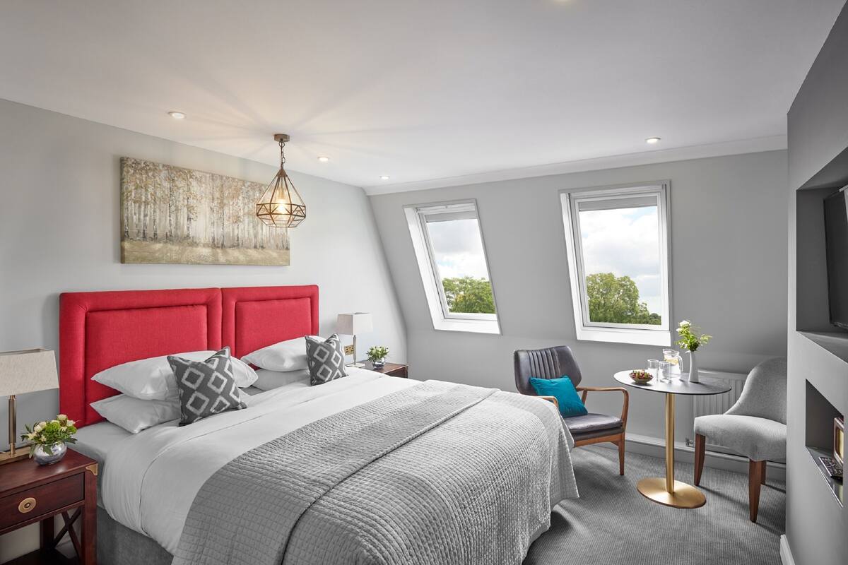 Quarto moderno com cabeceira vermelha, cama grande, mesa de cabeceira com abajur, mesinha redonda com um vaso e duas cadeiras.