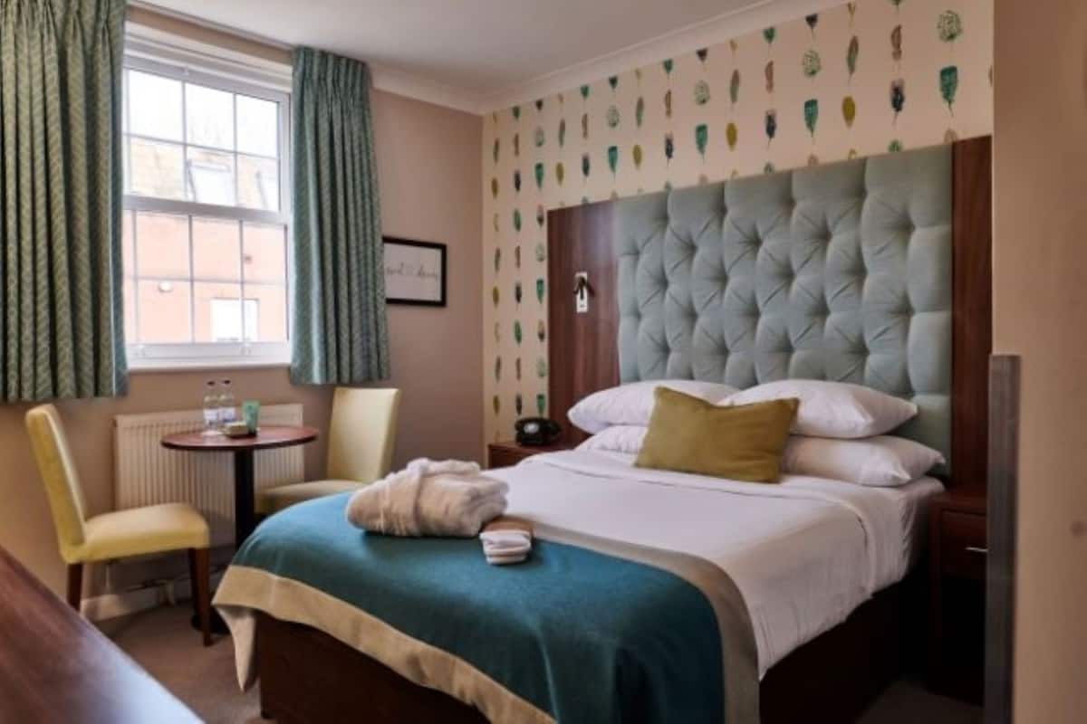 Quarto de hotel com uma cama grande, uma cadeira, uma mesinha e uma janela com cortinas.