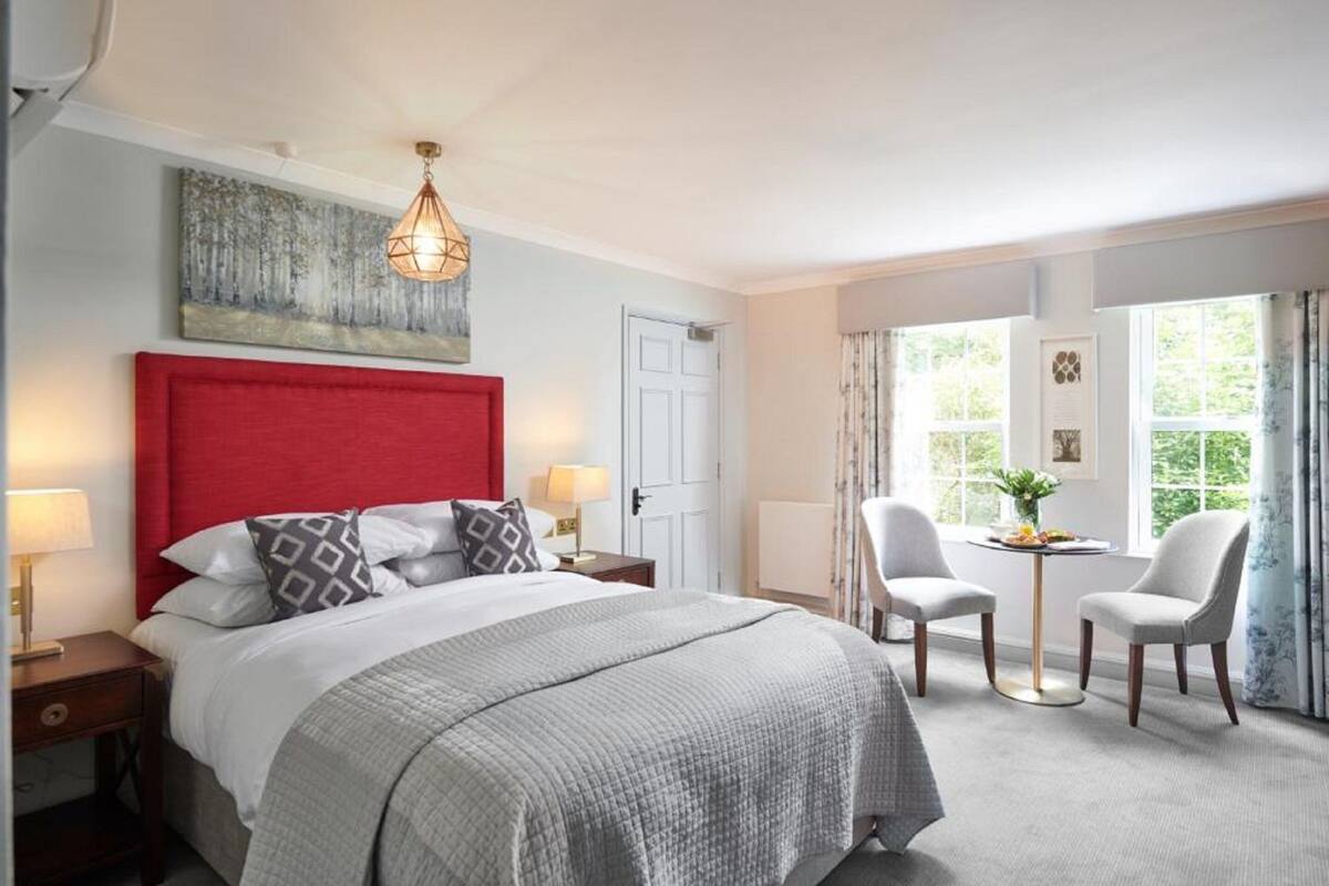 Quarto de hotel com cabeceira vermelha, cama com roupa de cama cinza, mesa de cabeceira com abajur, mesa redonda pequena com cadeiras e uma janela com cortinas.