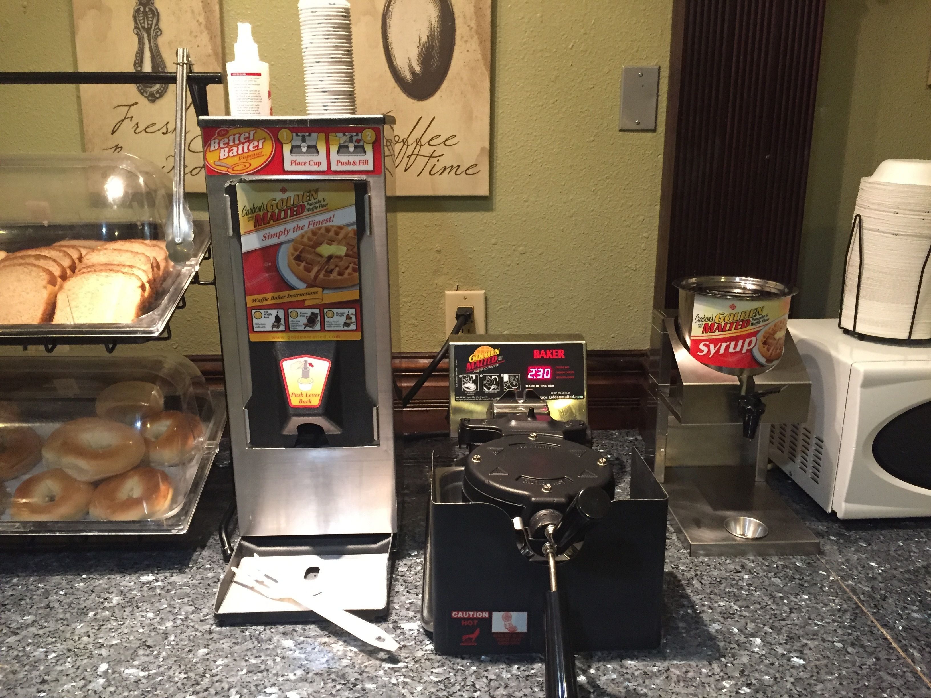 A coffee station with a waffle maker, syrup dispenser, and a display of bagels.