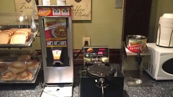 A coffee station with a waffle maker, syrup dispenser, and a display of bagels.