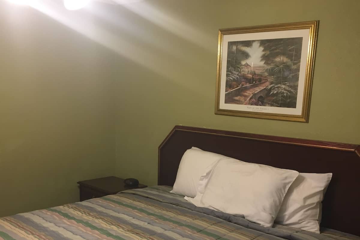 A hotel room with a bed, a nightstand, a framed picture on the wall, and a ceiling light fixture.