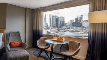 Une chambre d’hôtel avec vue sur la ville, un lit, deux fauteuils, une petite table avec un panier de fruits et une lampe.