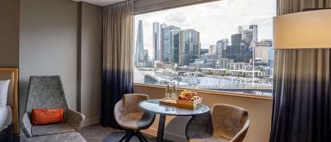 A hotel room with a city view, a bed, two armchairs, a small table with a fruit basket, and a lamp.
