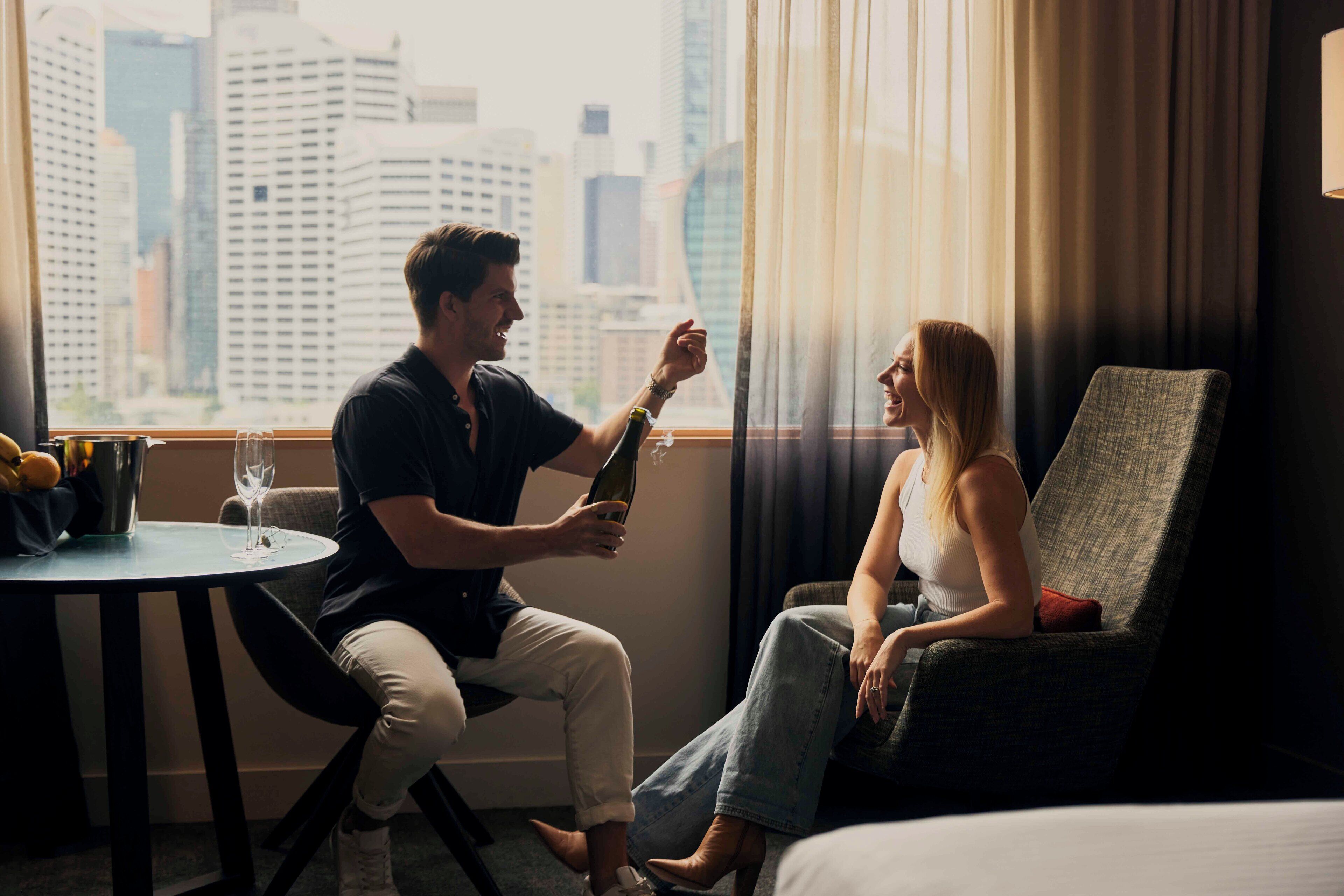A man and a woman sitting by a window with a city view, a table with a bottle and a glass, and a chair with a cushion.
