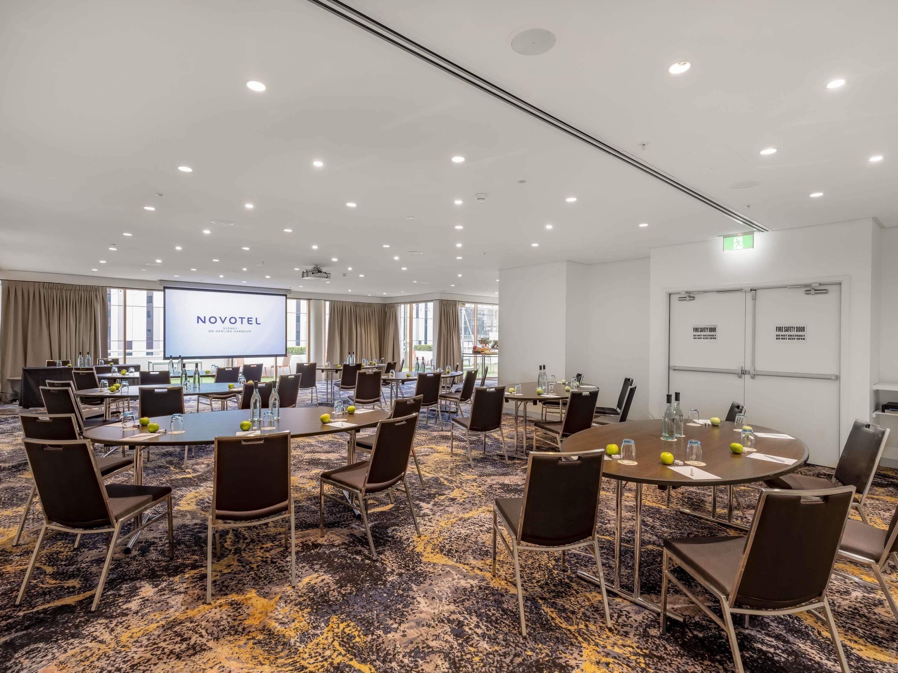 A conference room with multiple tables and chairs, a large screen displaying 'NOVOTEL,' and fire safety doors.