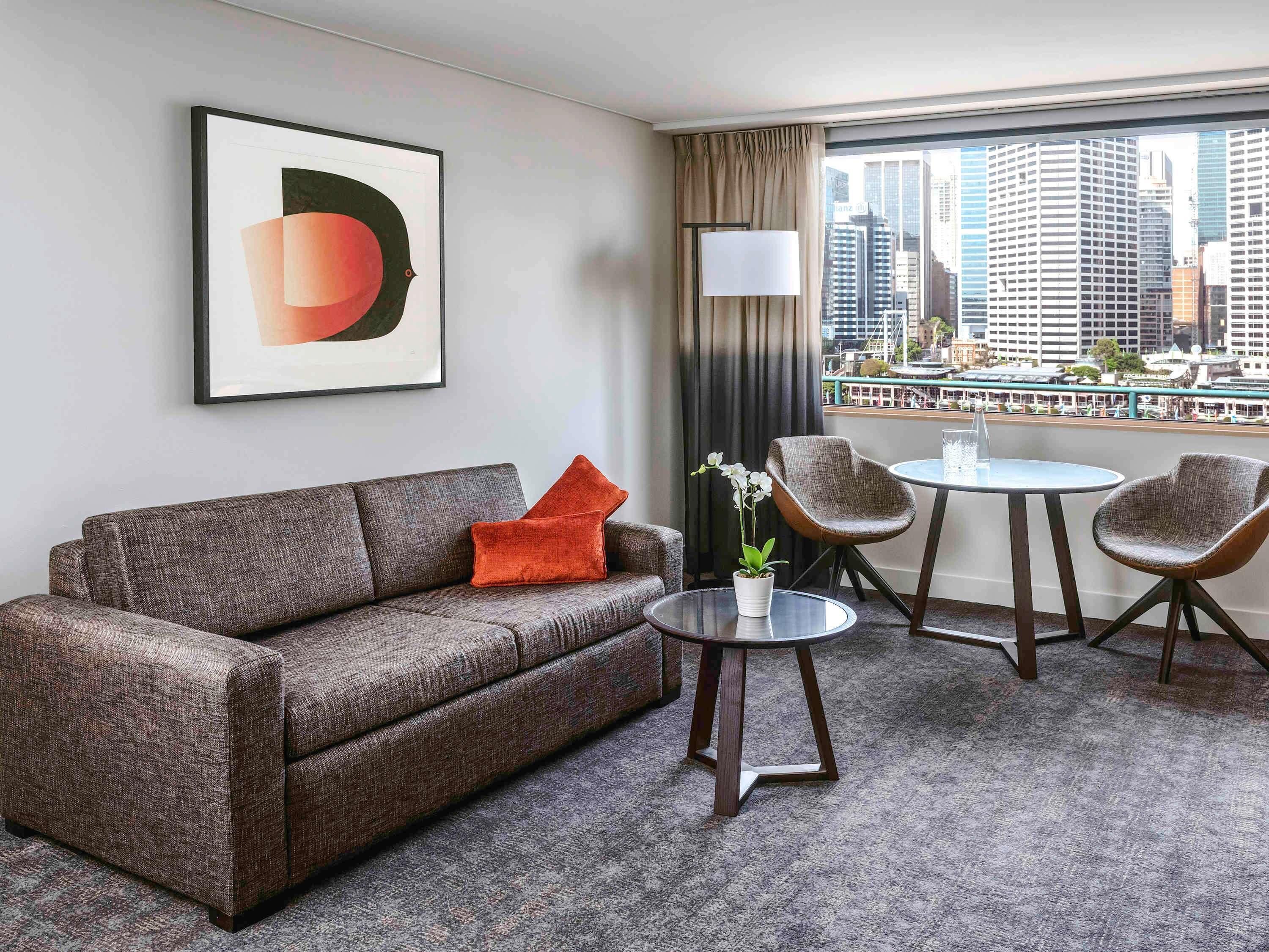 A hotel room with a sofa, two chairs, a small table, a potted plant, a lamp, and a city view through large windows.
