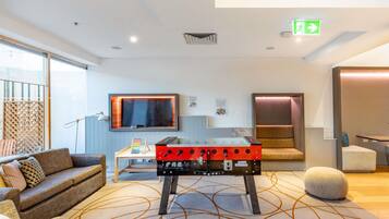 Une chambre d’hôtel moderne équipée d’un canapé, d’un baby-foot, d’un téléviseur fixé au mur et d’un tapis à motifs.