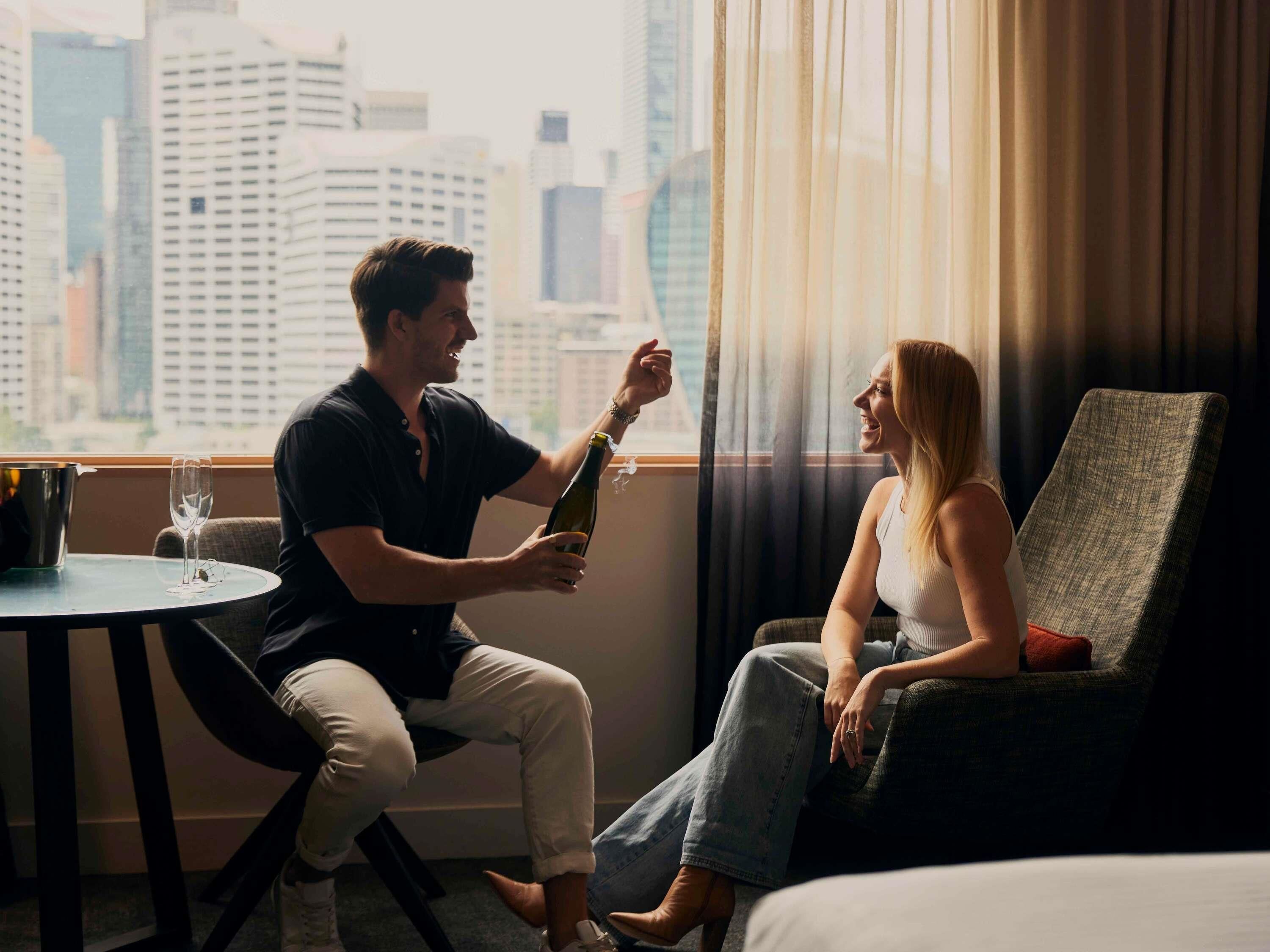 Ein Mann und eine Frau sitzen an einem Fenster mit Blick auf die Stadt, ein Tisch mit einer Flasche und einem Glas sowie ein Stuhl.