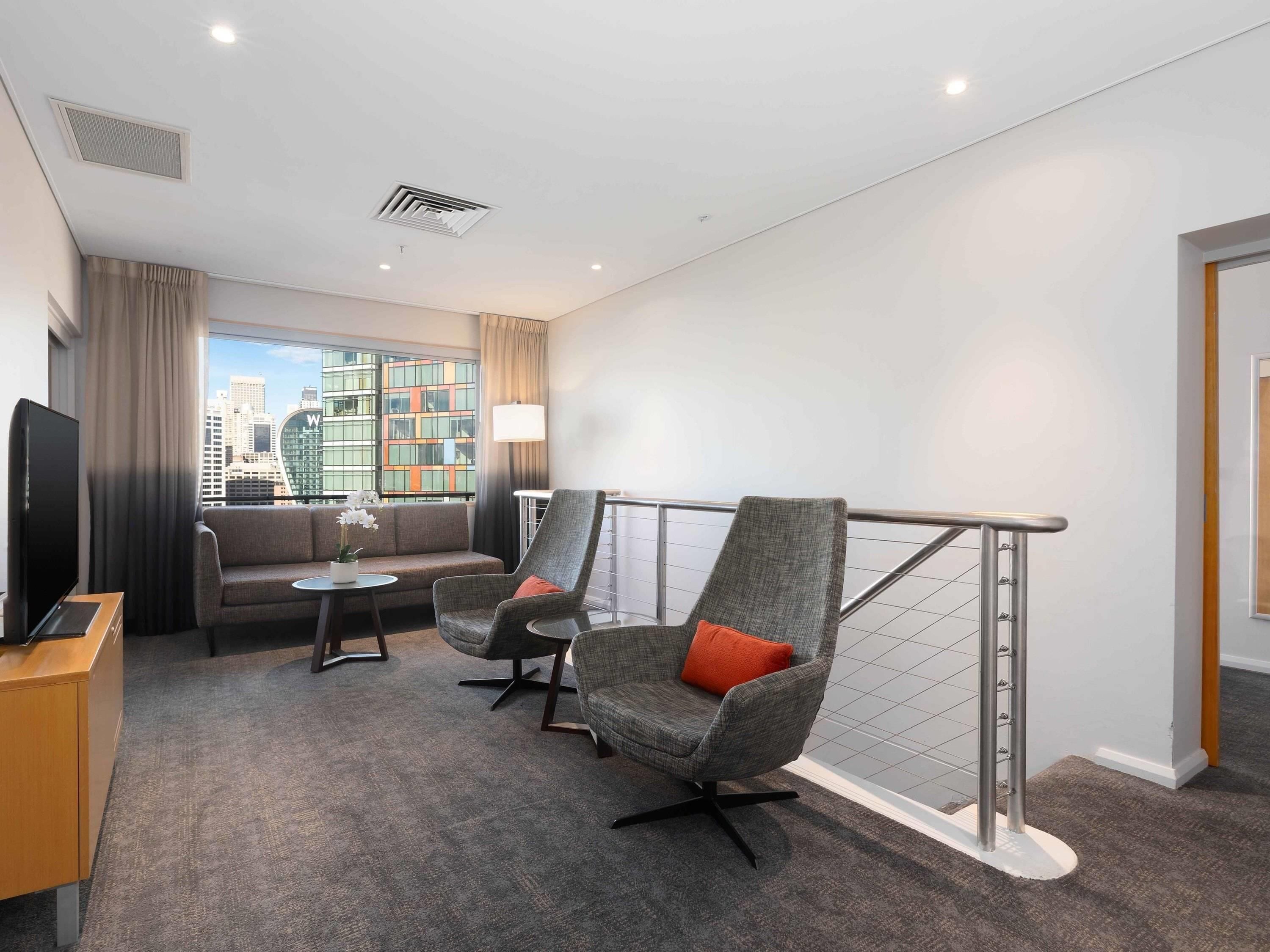 Habitación de hotel con sofá, dos sillones, televisor, una mesita y vistas a un paisaje urbano.