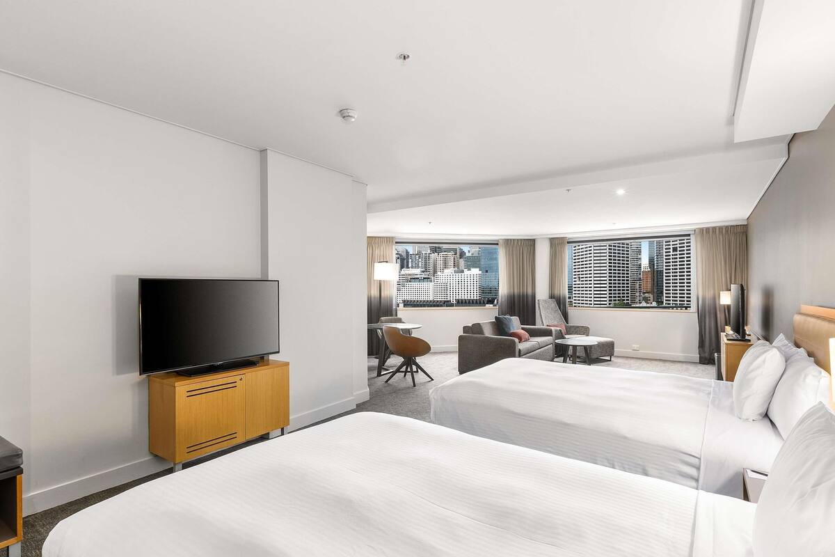 Ett hotellrum med två sängar, en tv-apparat placerad på ett träskåp, utsikt över staden genom stora fönster och en liten sittgrupp med ett bord och stolar.