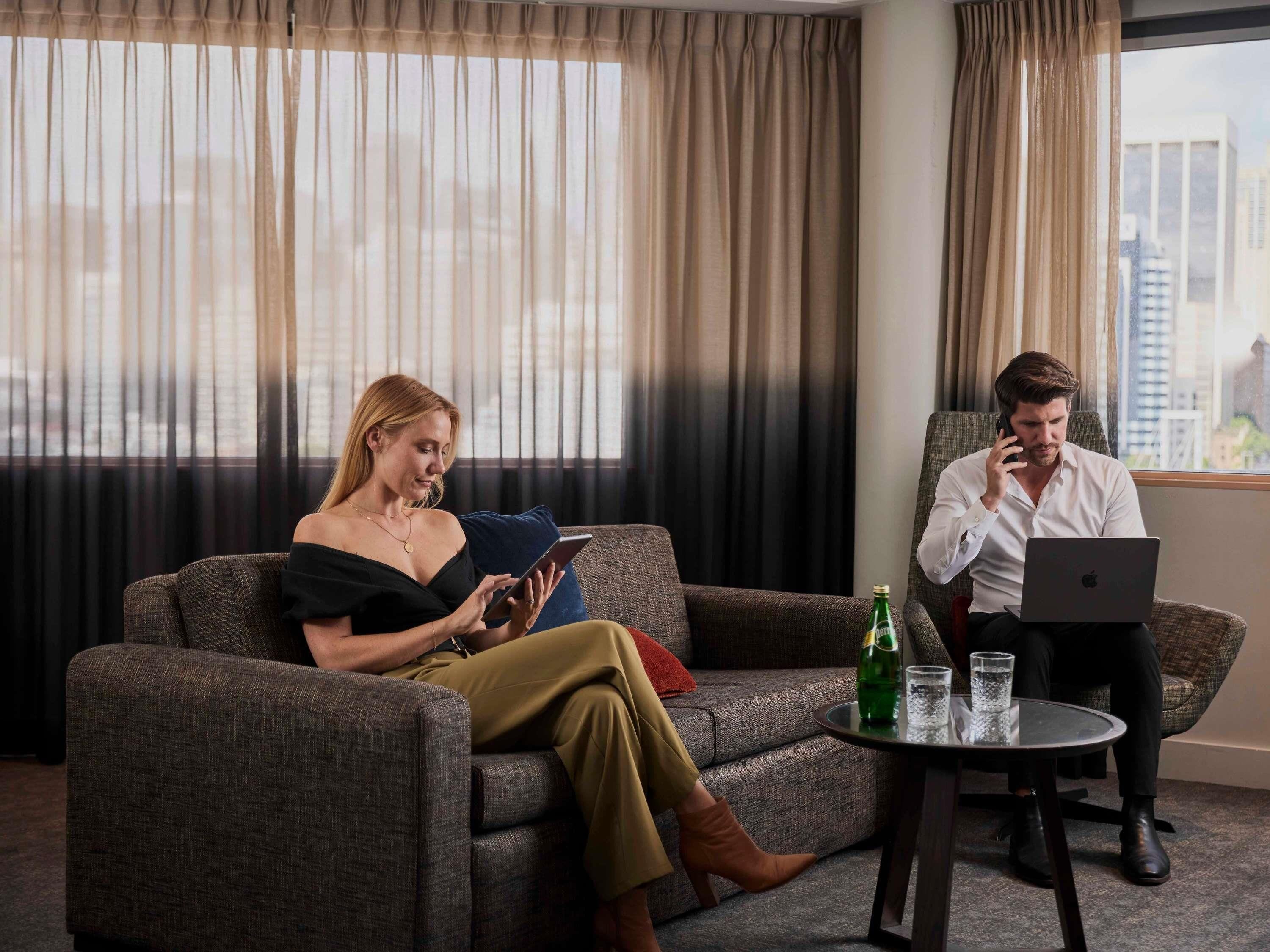 A modern hotel room with a sofa, a small table with a laptop, and a tablet.