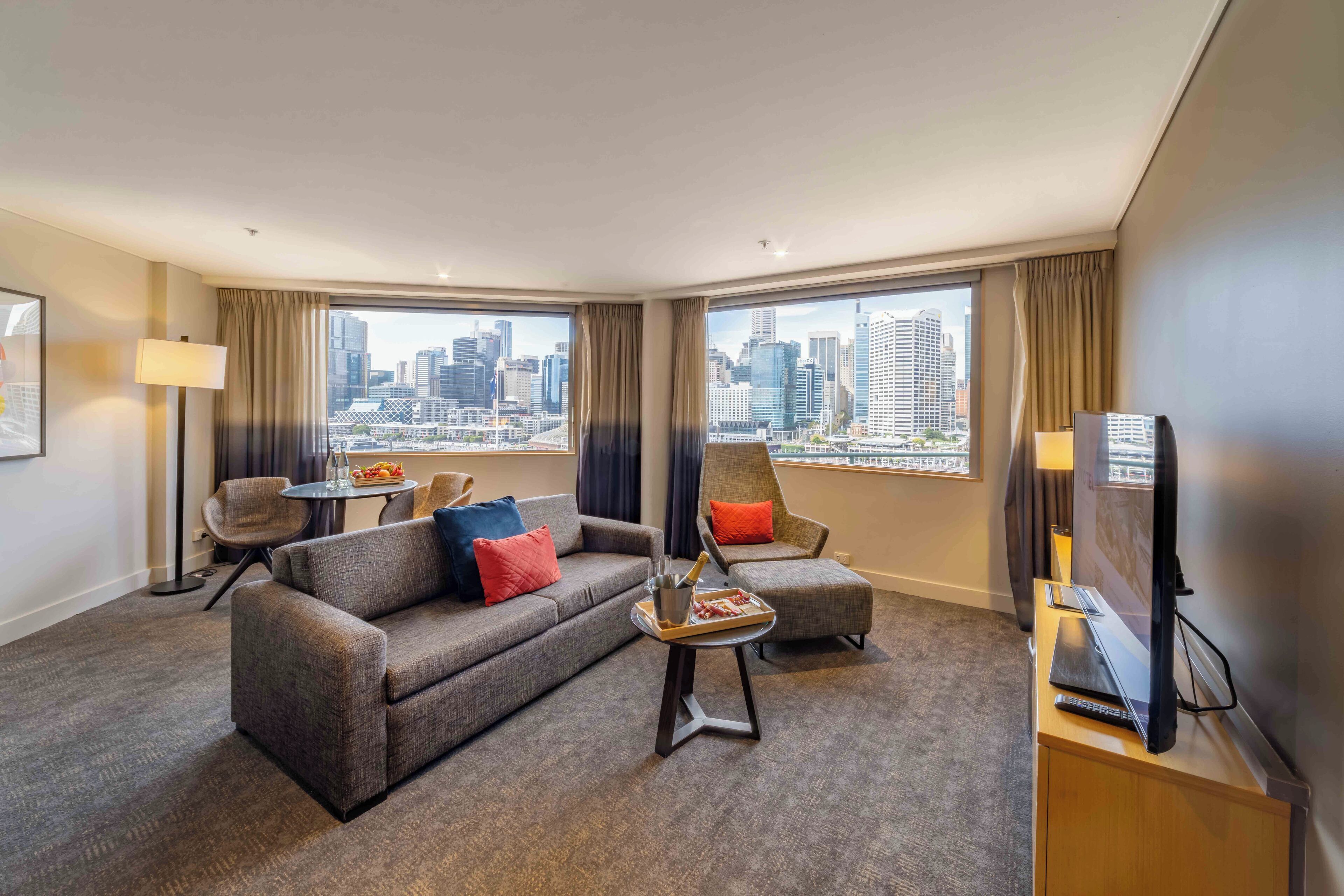 Een hotelkamer met een groot raam met uitzicht op de stad, een bank, een stoel, een klein tafeltje met dienblad en een televisie op een standaard.