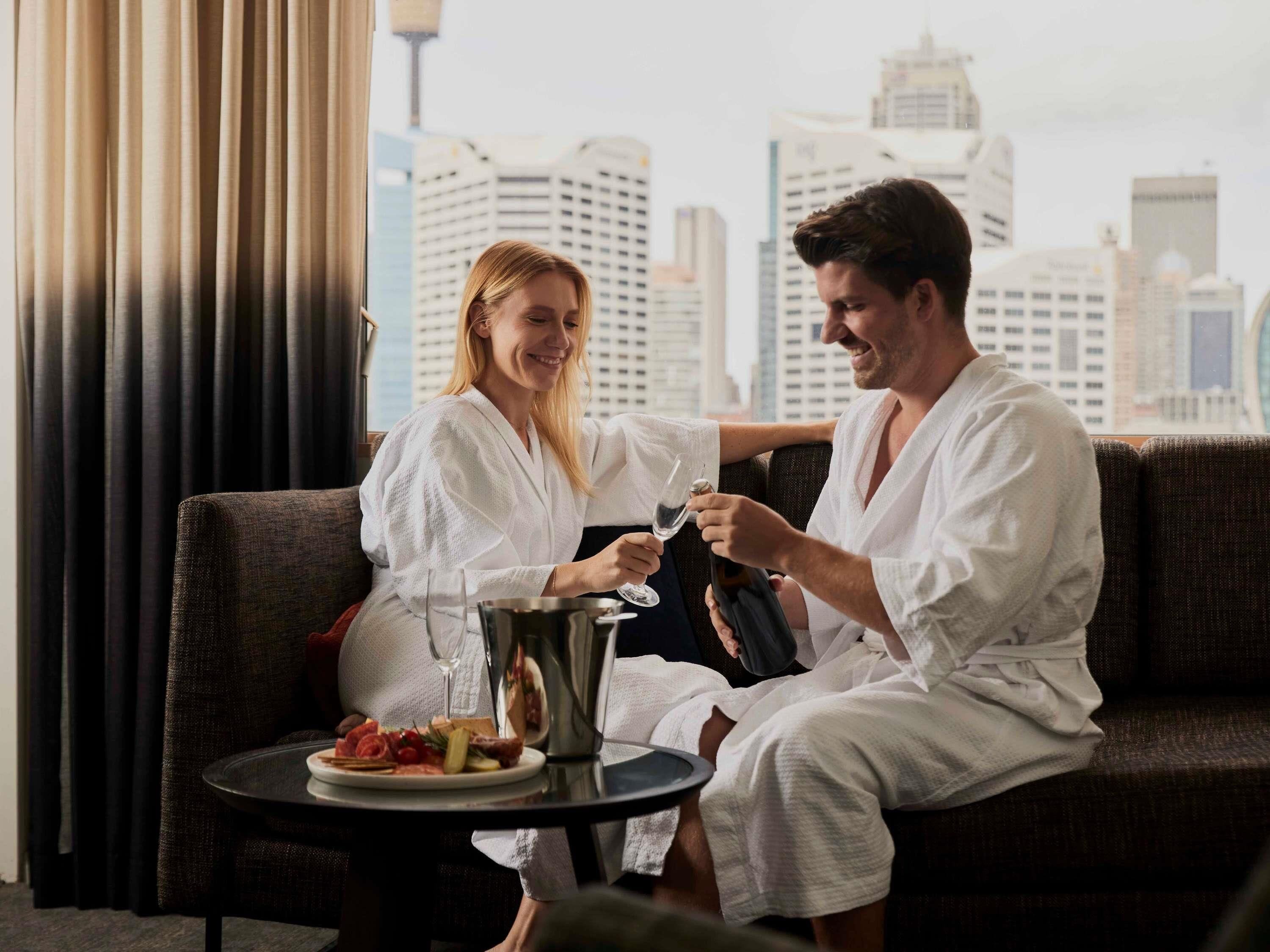 A couple in white robes sitting on a couch with a city view, a plate of food, and a wine glass on a small table.