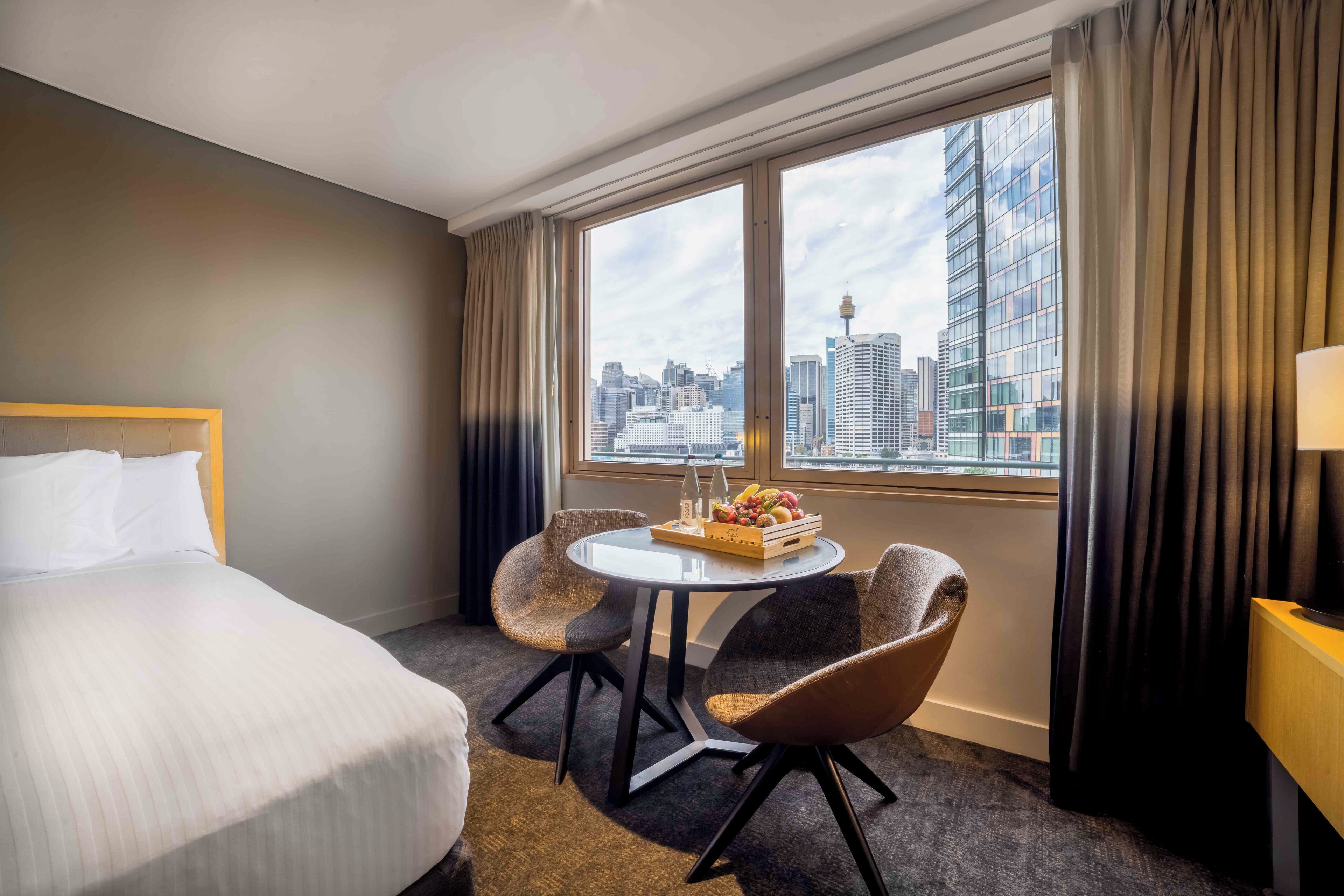 Quarto de hotel com uma cama, uma mesa redonda com cadeiras, vista para a cidade e uma luminária.
