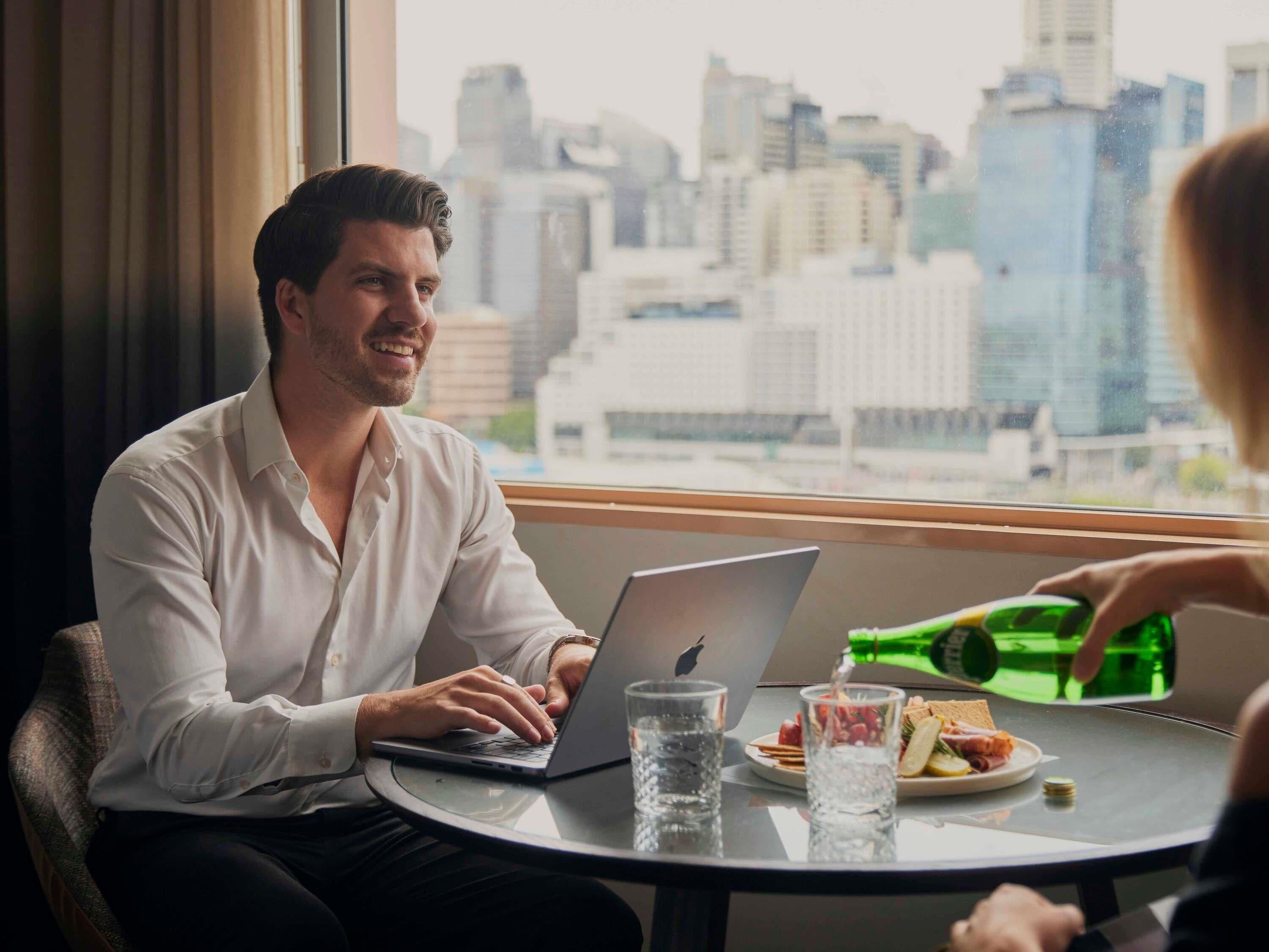 Una persona usando una computadora portátil en una mesa, con vista a la ciudad, un plato de comida y un vaso de agua.