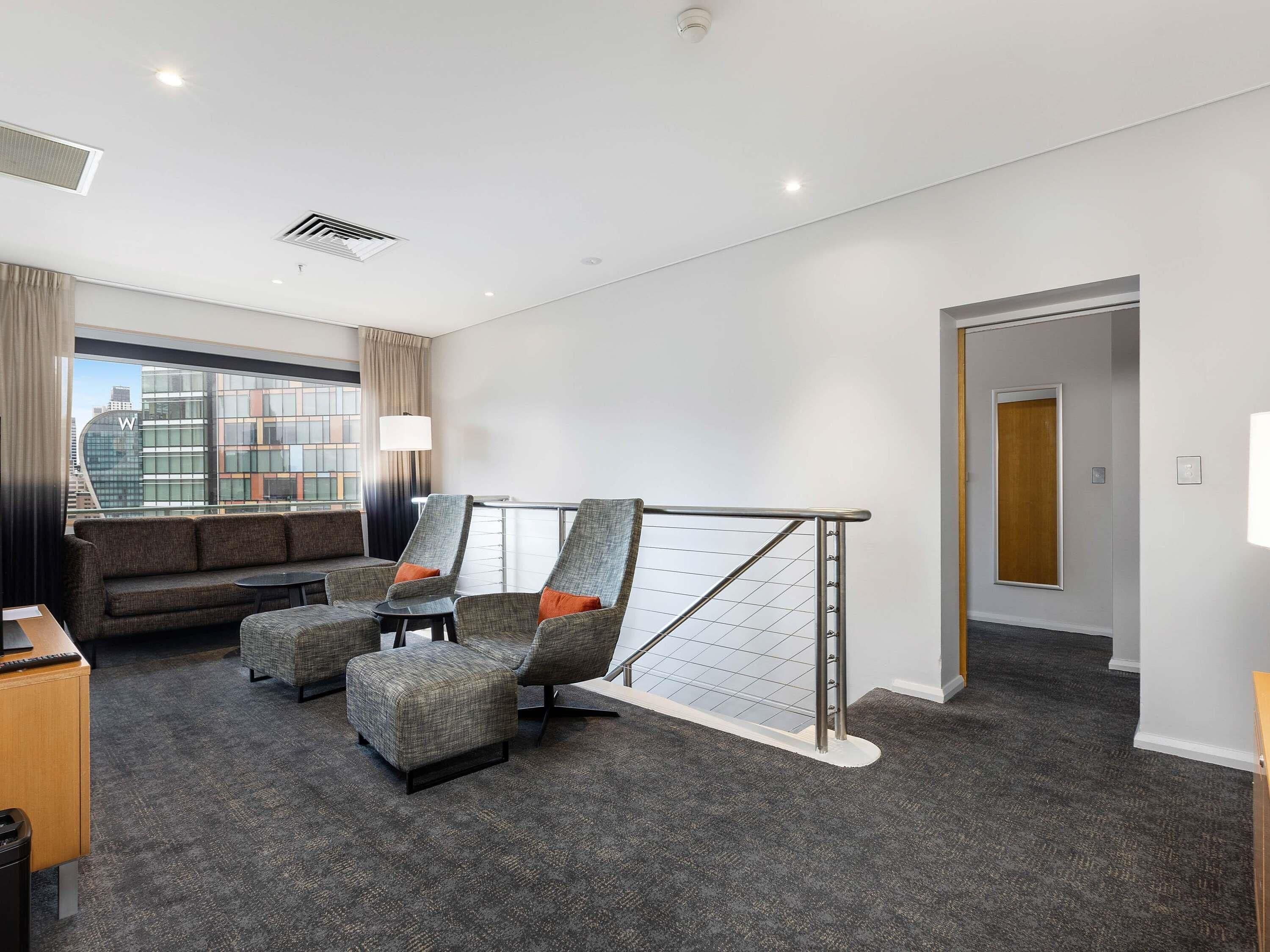 A hotel room with a sofa, two armchairs, a small table, a window with a view of buildings, and a door.