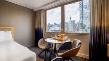 Habitación de hotel con una cama, dos sillones, una mesita con fruta y vistas a la ciudad a través de amplias ventanas.