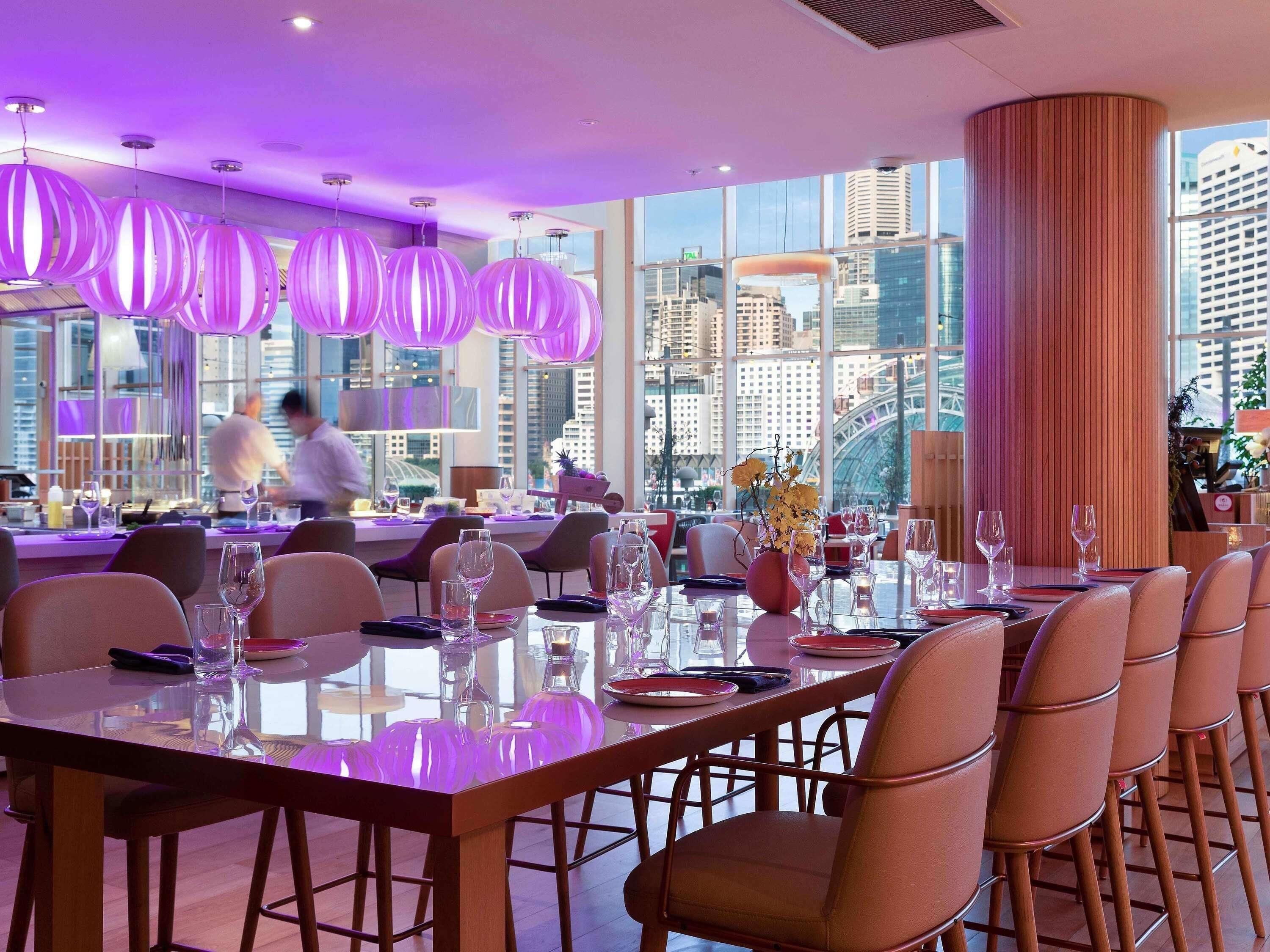 Un restaurante moderno con una larga mesa de comedor, lámparas colgantes púrpuras y vistas a la ciudad.