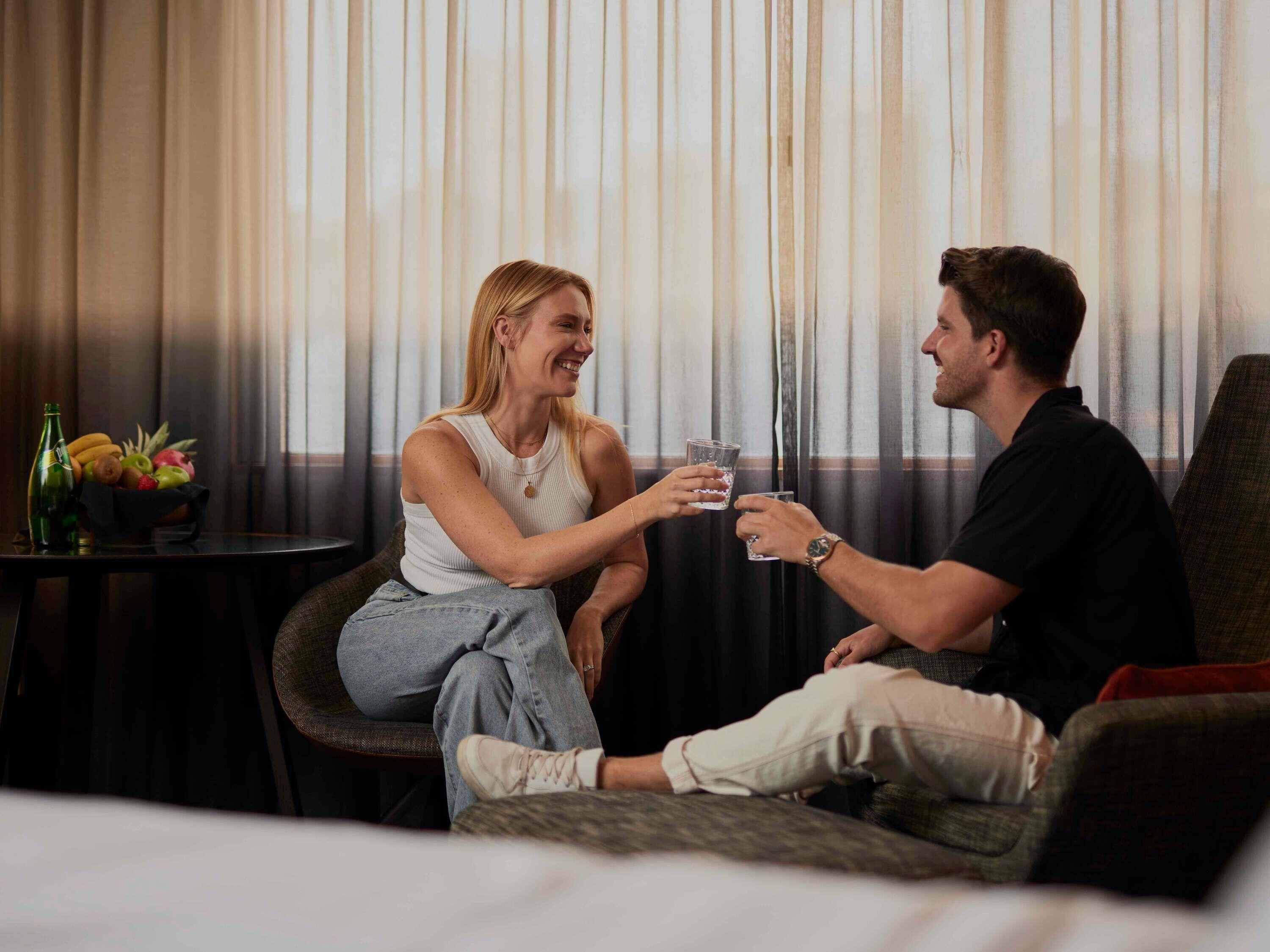 Un couple est assis sur un canapé, tenant des verres à la main, avec une table remplie de fruits et une bouteille en arrière-plan.