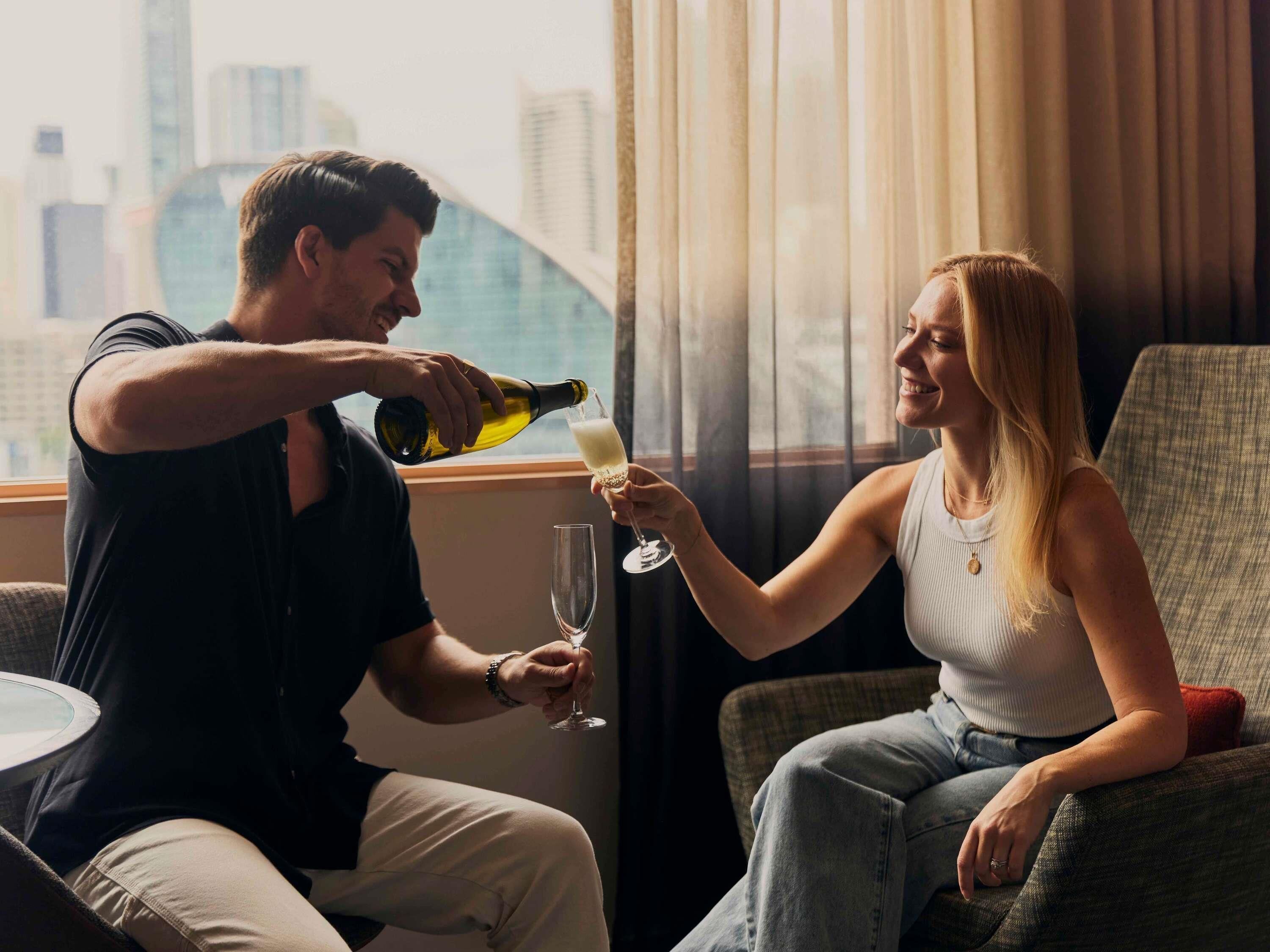 Un hombre sirve una bebida en un vaso que sostiene una mujer, ambos sentados junto a una ventana con vistas a la ciudad.
