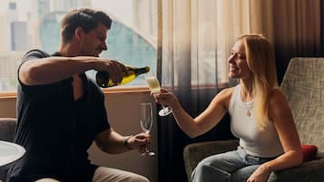 Un hombre sirve una bebida en un vaso que sostiene una mujer, ambos sentados junto a una ventana con vistas a la ciudad.