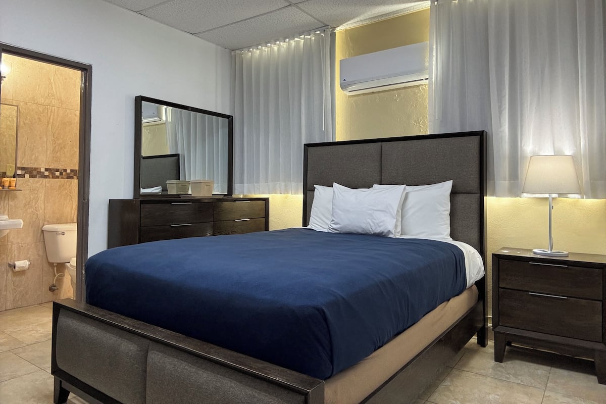 A hotel room with a bed, a nightstand, a mirror, a toilet, and an air conditioning unit.