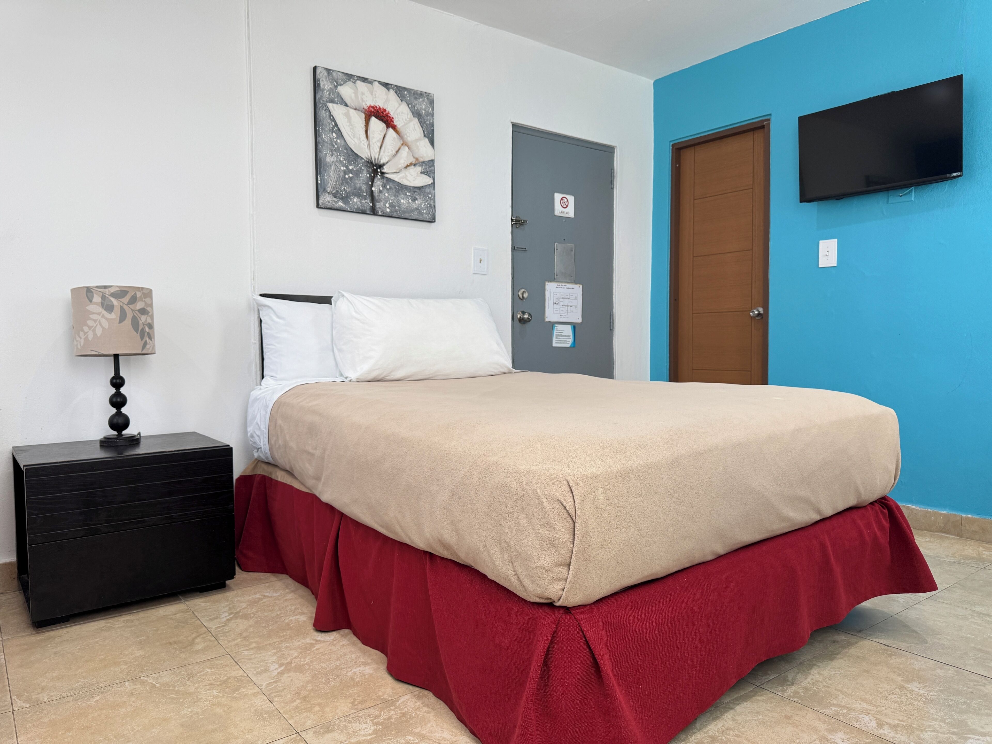 A bedroom with a bed, a nightstand, a lamp, a painting, a door, and a TV.