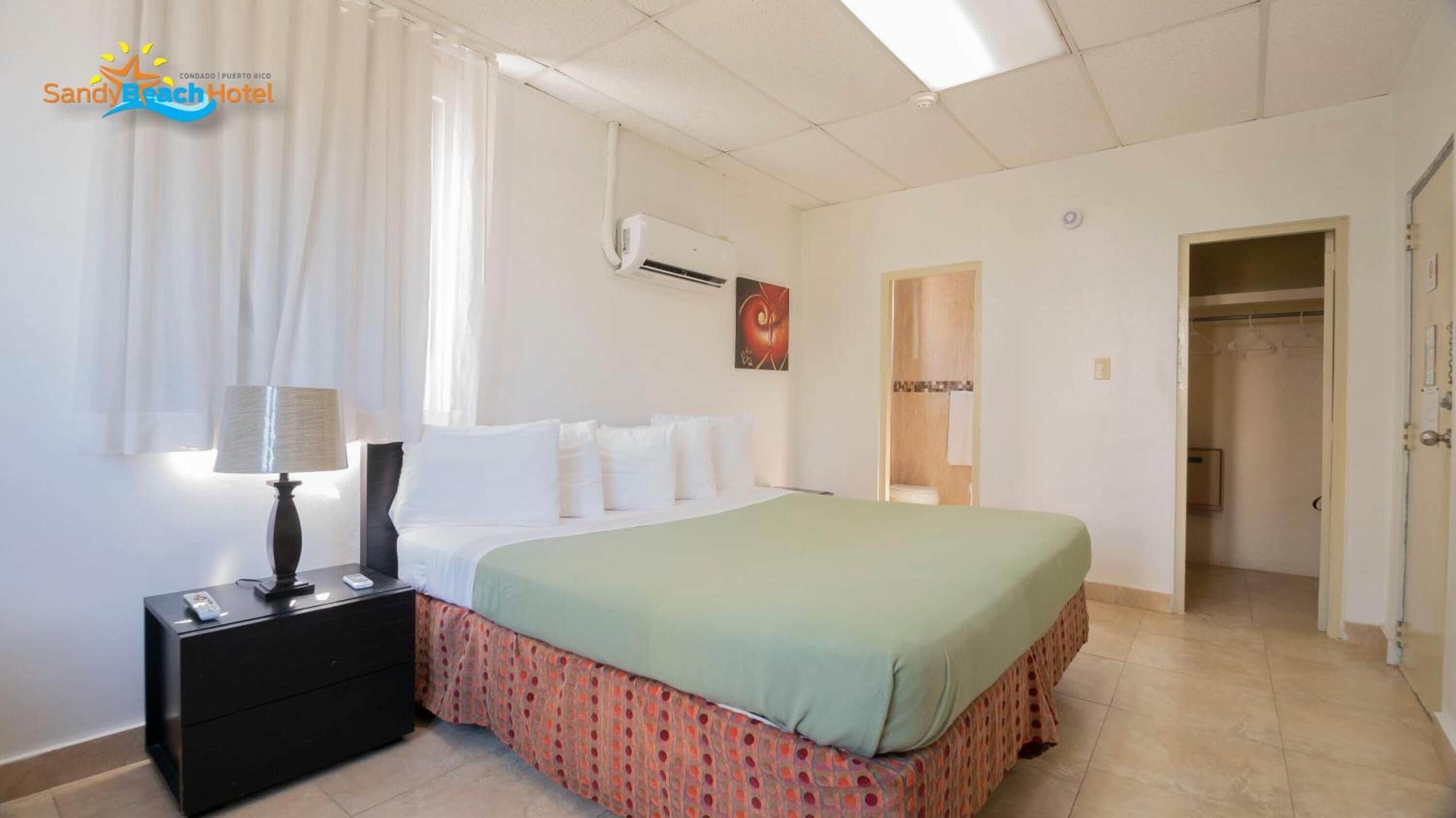 Una habitación de hotel con cama, mesita de noche, aire acondicionado, un cuadro y la puerta del baño.
