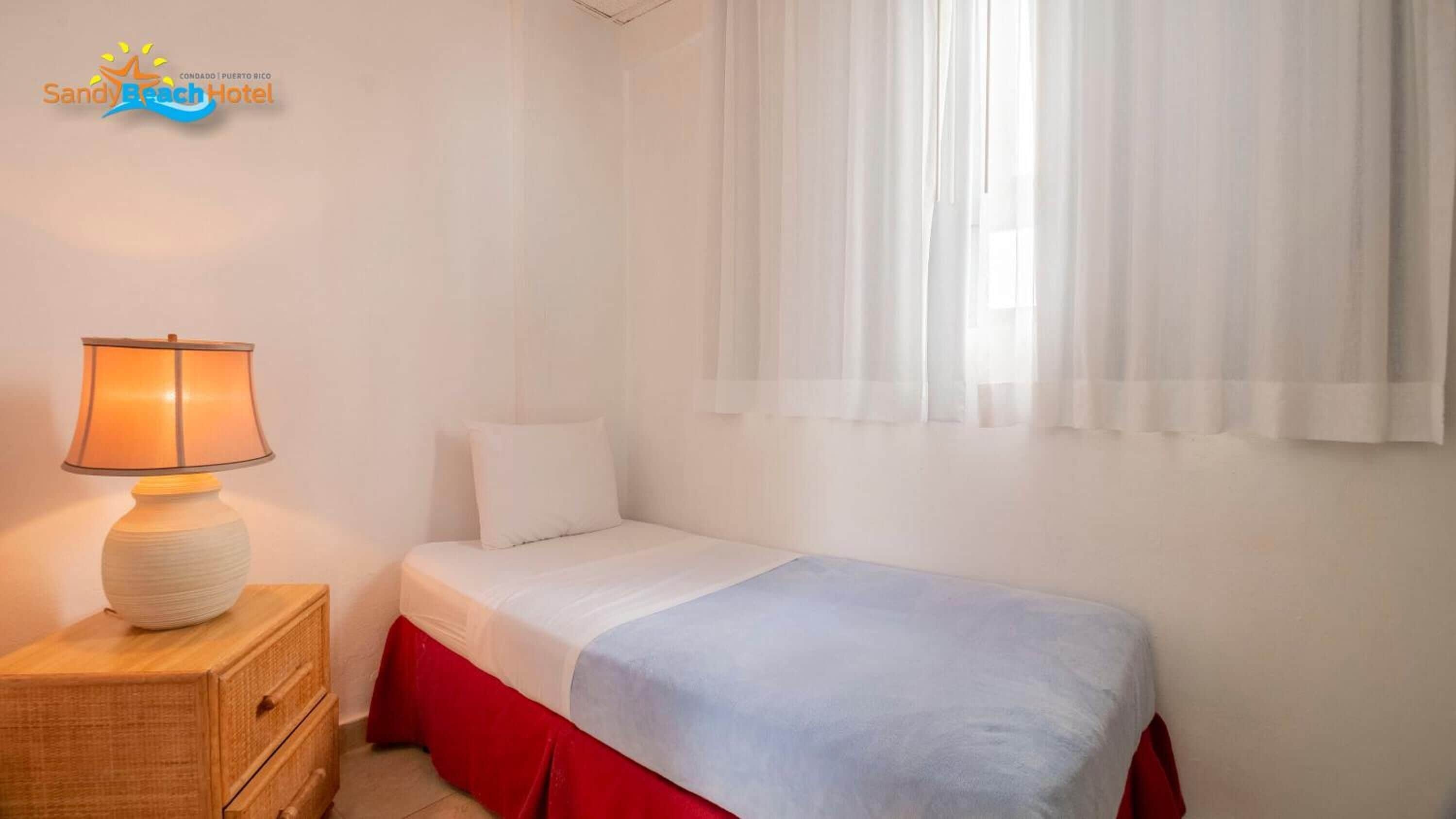 A simple bedroom with a single bed, a bedside lamp, and a window with sheer curtains.