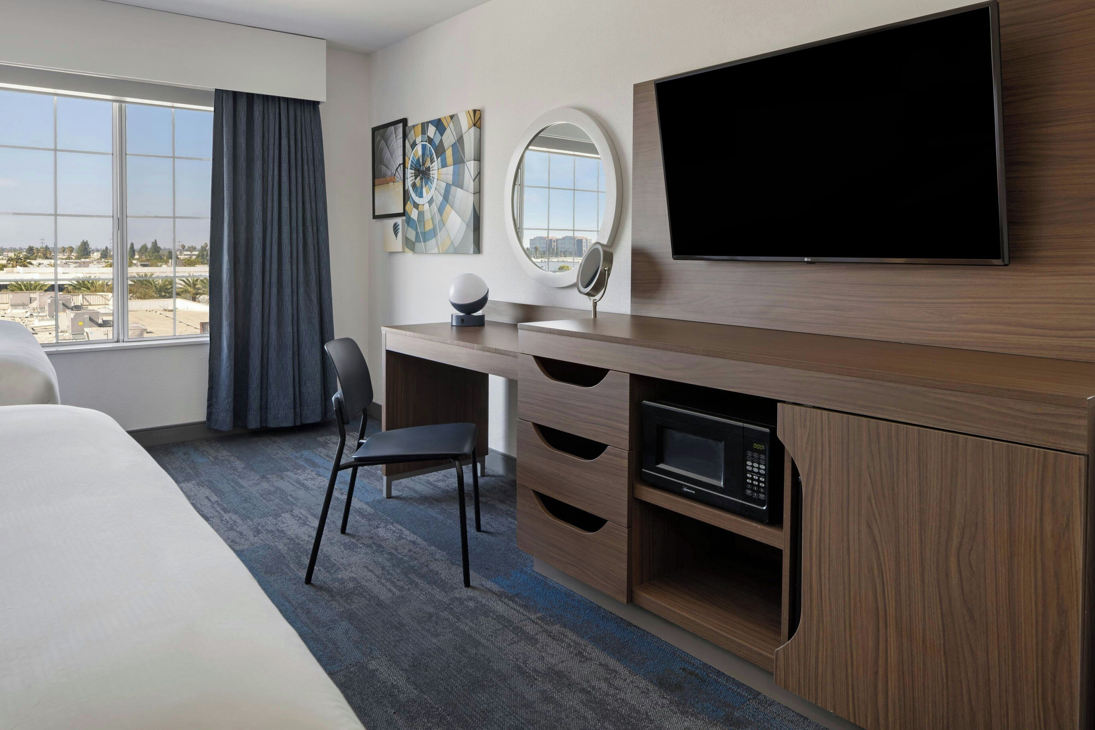 Habitación de hotel con una cama grande, un escritorio, un televisor de pantalla plana, un microondas y vistas a la ciudad desde un ventanal amplio.
