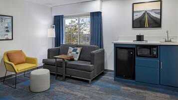 A modern hotel room with a grey sofa, a yellow armchair, a small table, a white ottoman, a kitchen area with a microwave, and a large window with blue curtains.