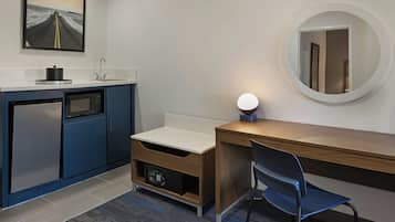 Habitación compacta con kitchenette, escritorio de madera, una silla, un espejo redondo y un cuadro en la pared.