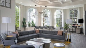 A modern living room with a sectional sofa, a coffee table, and a TV. There are large windows with curtains, a plant, and a desk with a chair. - City Express by Marriott Santa Ana/Orange County Airport (Santa Ana)