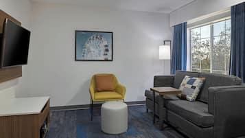 A modern hotel room with a grey sofa, a yellow armchair, a small table, a TV mounted on the wall, and a large window with curtains.
